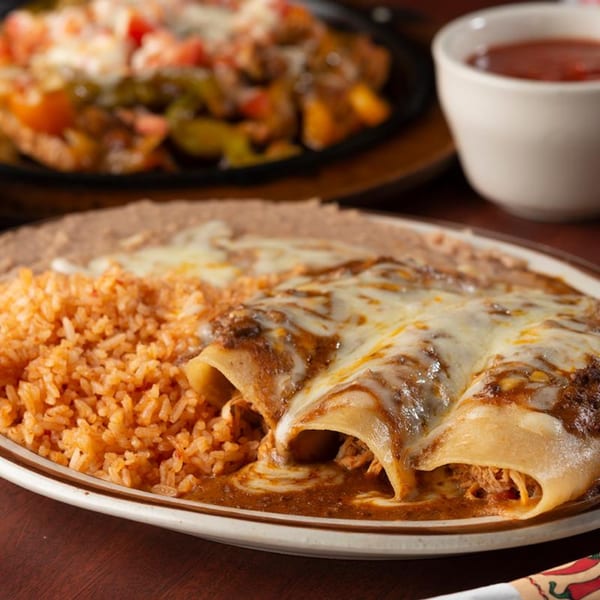 Three cheesy enchiladas with rice and refried beans, with sizzling fajitas in the background.