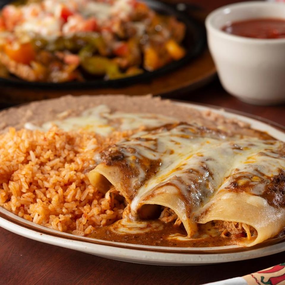 Three cheesy enchiladas with rice and refried beans, with sizzling fajitas in the background.